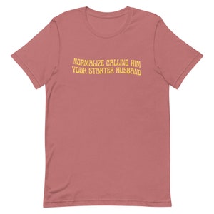 May include: A dusty rose-colored t-shirt with the text "NORMALIZE CALLING HIM YOUR STARTER HUSBAND" in a yellow, slightly distressed font. The shirt has short sleeves and a crew neck.