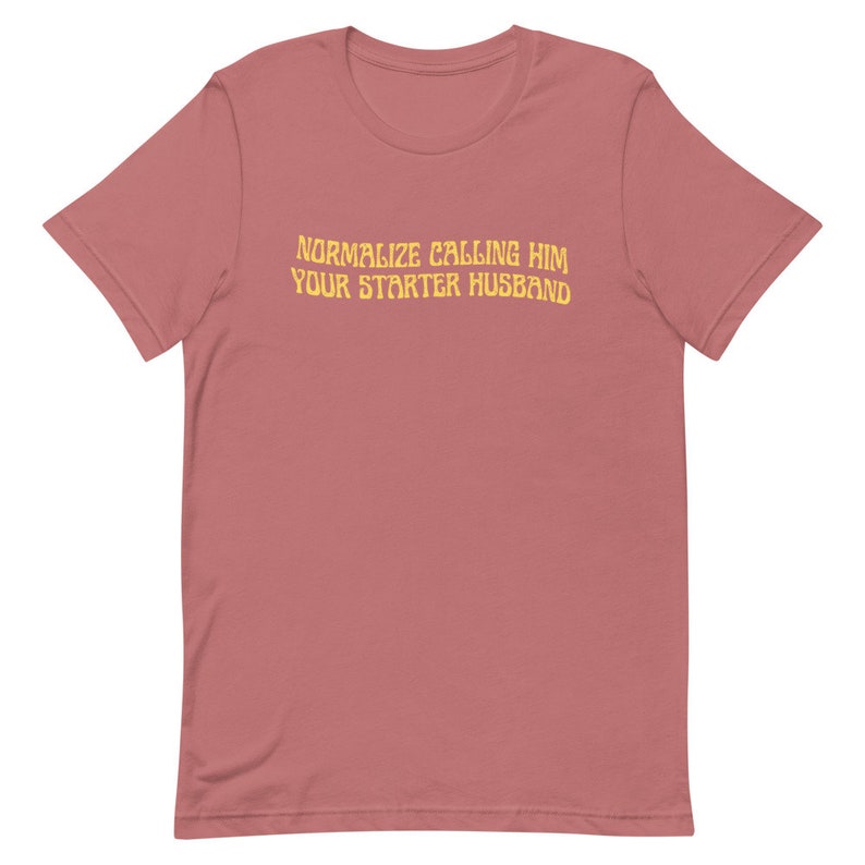 May include: A dusty rose-colored t-shirt with the text "NORMALIZE CALLING HIM YOUR STARTER HUSBAND" in a yellow, slightly distressed font. The shirt has short sleeves and a crew neck.
