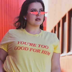 May include: A woman wearing a yellow t-shirt with the text "YOU'RE TOO GOOD FOR HIM" in red lettering. She is wearing a red belt and blue jeans. She is also wearing red flame-shaped sunglasses.
