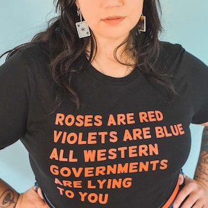 Könnte beinhalten: Schwarzes T-Shirt mit dem orangefarbenen Text "Roses are red, violets are blue, all western governments are lying to you." Die Person trägt Spielkartenohrringe. Der Hintergrund ist hellblau.