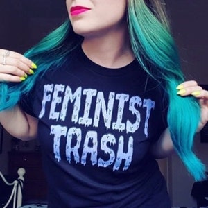 May include: Black t-shirt with the text "FEMINIST TRASH" in white and blue lettering. The person in the image has bright teal hair and is wearing bright pink lipstick.