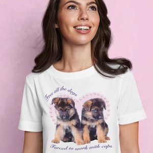 Può includere: T-shirt bianca con due cuccioli di pastore tedesco all'interno di un cuore rosa. La maglietta presenta la scritta "Free all the dogs" e "Forced to work with cops" in corsivo.