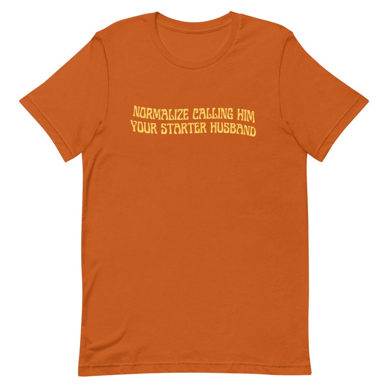 May include: An orange t-shirt with the text "NORMALIZE CALLING HIM YOUR STARTER HUSBAND" in a yellow, retro font. The shirt is a solid color and appears to be made of a soft material.