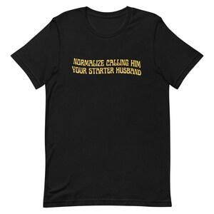 May include: Black t-shirt with the text "NORMALIZE CALLING HIM YOUR STARTER HUSBAND" in a gold, slightly distressed font. The shirt has a crew neck and short sleeves.