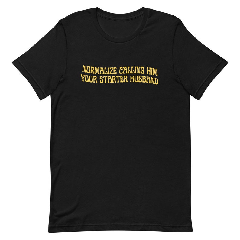 May include: Black t-shirt with the text "NORMALIZE CALLING HIM YOUR STARTER HUSBAND" in a gold, slightly distressed font. The shirt has a crew neck and short sleeves.