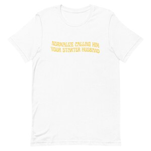 May include: White t-shirt with the text "NORMALIZE CALLING HIM YOUR STARTER HUSBAND" in a gold color. The shirt is made of a soft material and has short sleeves. The text is centered on the front of the shirt.