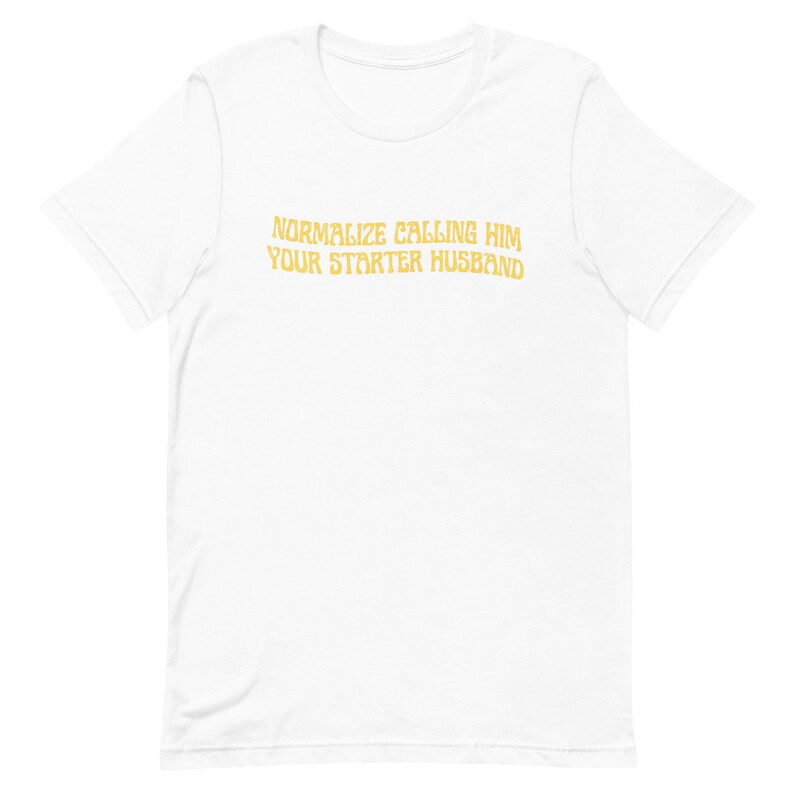 May include: White t-shirt with the text "NORMALIZE CALLING HIM YOUR STARTER HUSBAND" in a gold color. The shirt is made of a soft material and has short sleeves. The text is centered on the front of the shirt.
