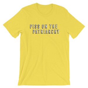 Puede incluir: Camiseta amarilla de manga corta con el texto "PISS ON THE PATRIARCHY" impreso en negro.