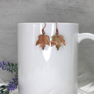 May include: A pair of copper and gold leaf earrings in the shape of maple leaves. The earrings are hanging from a white mug.