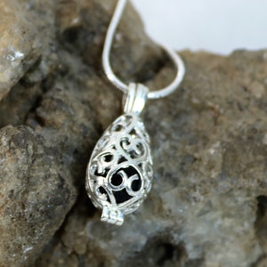 May include: A silver filigree pendant necklace with a teardrop-shaped cage. The cage is open and has a black stone inside. The necklace is on a rock.