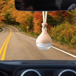 Puede incluir: Un ambientador de coche de madera colgado del espejo retrovisor de un coche. El coche está conduciendo por una carretera con follaje de otoño a ambos lados.