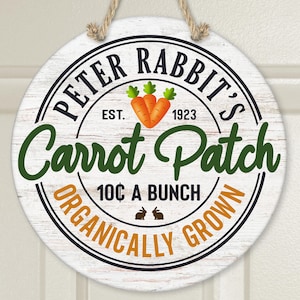 Puede incluir: Un letrero redondo blanco con letras negras y verdes que dice "Peter Rabbit's Carrot Patch Est. 1923 10¢ A Bunch Organically Grown". El letrero tiene un fondo de madera rústica y presenta una imagen de dibujos animados de dos zanahorias.