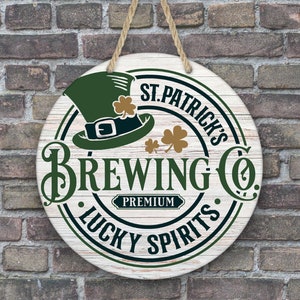 Puede incluir: Letrero redondo de madera con el texto "St. Patrick's Brewing Co. Lucky Spirits Premium" en verde y negro. El letrero presenta un sombrero de duende verde y tréboles. Cuelga de una cuerda natural y está montado en una pared de ladrillo.
