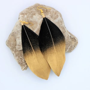 May include: A pair of long feather earrings with a black to gold ombre effect. The feathers are black at the top and fade to gold at the bottom.