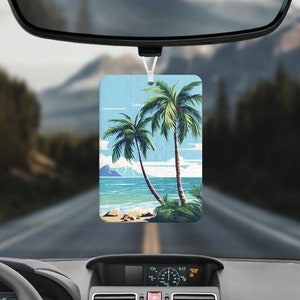 May include: Rectangular air freshener featuring a tropical beach scene with two palm trees, blue water, and a light blue sky. The air freshener is hanging from a car's rearview mirror, with a blurred background of a road and mountains.