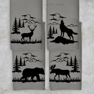 Puede incluir: Conjunto de cuatro toallas de cocina grises con diseños de silueta negra de un ciervo, un lobo, un oso y un alce en un entorno de bosque con montañas y luna llena. El texto "BETS.COM" está impreso en una de las toallas.