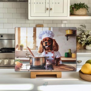 Op de afbeelding: Een bruine en witte Cavalier King Charles Spaniel hond die een witte koksmuts en een rood-witte koksjas draagt, staat in een keukensituatie. De hond kijkt naar de camera en heeft een pan met kokend water op het fornuis achter hem. De hond staat op een houten snijplank. De afbeelding is een keuken-thema ontwerp voor een snijplank.