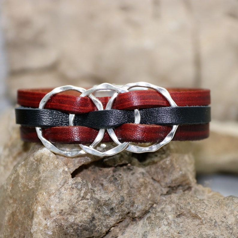 Op de afbeelding: Een leren armband met drie zilveren ringen die in de zwarte en bruine leren banden zijn geweven.