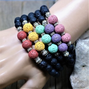 Puede incluir: Varios brazaletes con cuentas de roca volcánica negra y cuentas porosas de colores rojo, amarillo, verde, morado y rosa. Los espaciadores plateados separan las cuentas. Los brazaletes se muestran en el brazo de una persona.