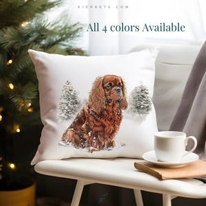 Puede incluir: Cojín cuadrado blanco con un Cavalier King Charles Spaniel en una escena de bosque nevado. El perro es marrón y canela. El texto "All 4 colors Available" es visible. También hay una taza y un platillo blancos.