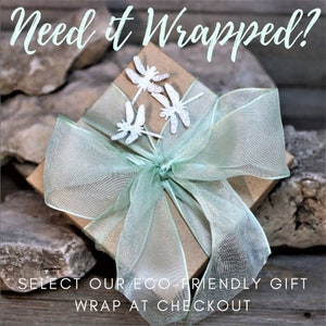 Op de afbeelding: Een bruine cadeaudoos verpakt in een lichtgroen, doorschijnend lint met zilveren libellenversieringen. De doos staat op een rustiek houten oppervlak. "Need it Wrapped?" staat in witte tekst bovenaan de afbeelding. "Select our eco-friendly gift wrap at checkout" staat in zwarte tekst onderaan de afbeelding.