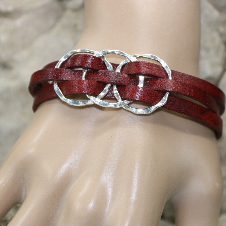 Op de afbeelding: Een bruin leren armband met drie zilveren ringen die in het leer zijn geweven. De ringen zijn gehamerd en hebben een onregelmatige vorm.