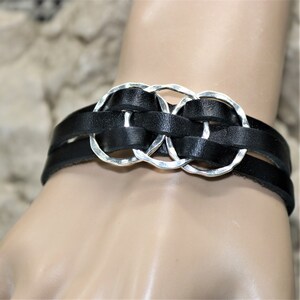 Op de afbeelding: Een zwart leren armband met drie zilveren, in elkaar grijpende cirkels. De armband is geweven en heeft een eenvoudig ontwerp.