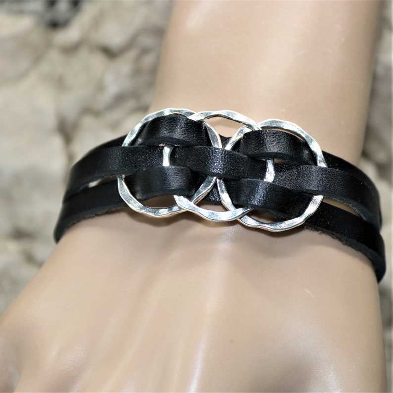 Op de afbeelding: Een zwart leren armband met drie zilveren, in elkaar grijpende cirkels. De armband is geweven en heeft een eenvoudig ontwerp.