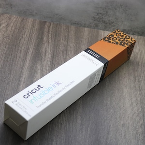 May include: A white box with the Cricut logo and the text "infusible ink" in purple. The box says "Transfer Sheets/Feuilles de transfert" and "2 sheets/20.3 cm x 30.5 cm". The box is on a brown wooden surface. The top of the box has a leopard print design.