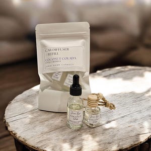 May include: A car air freshener refill set with a white bag labeled "CAR DIFFUSER REFILL COCONUT COLADA". Includes a glass bottle with a dropper, a small bottle with a wooden top, and refill pads. The set is on a wooden surface.