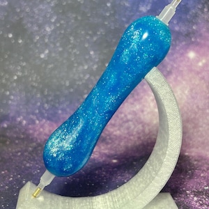 Puede incluir: Un bolígrafo de resina azul brillante con purpurina para diamond painting, con un cuerpo grueso y ergonómico. El bolígrafo se exhibe sobre un soporte blanco curvo y translúcido que presenta el texto verde en relieve "Lassen Lathe Works".