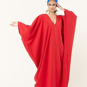 May include: A woman wearing a red kaftan with a blue head wrap and gold earrings. The kaftan has a V-neck and long sleeves.