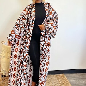 May include: A long white and brown patterned kimono with geometric designs. The kimono has long sleeves and a wide open front. The model is wearing a black mock neck shirt and black heels.