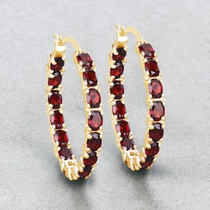 May include: A pair of gold-colored hoop earrings. Each earring is adorned with a row of oval-shaped, deep red gemstones. The earrings are displayed against a neutral gray background, highlighting the jewelry's design and color.