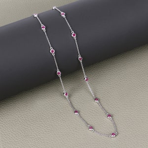 May include: A silver chain necklace with small red gemstones. The necklace is on a gray textured surface.