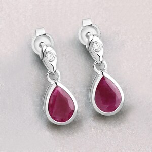 May include: Silver drop earrings with pear-shaped, deep red gemstones. Each earring has a small, clear gemstone accent at the top and a post back. The earrings are displayed against a light grey background.