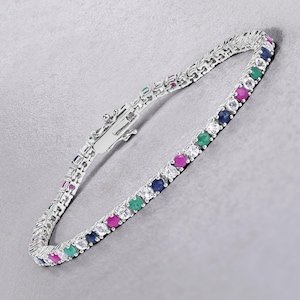 Puede incluir: Una pulsera de tenis plateada con cierre, con una fila de piedras preciosas alternas. Las piedras preciosas incluyen piedras blancas, azules, verdes y rosas. La pulsera se muestra sobre una superficie gris claro.