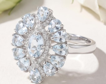 May include: A silver ring featuring a cluster of light blue gemstones. The ring's centerpiece is an oval-shaped stone, surrounded by smaller, pear-shaped stones and accent diamonds. The ring's band is a simple, polished silver.