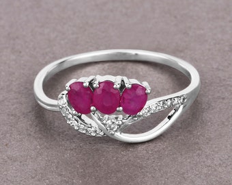 Puede incluir: Un anillo de plata con tres piedras preciosas de rubí de forma ovalada y piedras preciosas más pequeñas de color claro engastadas en un diseño delicado y curvo.