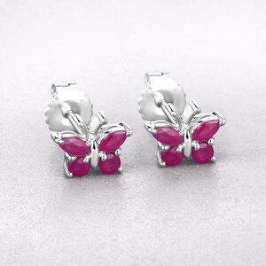 May include: A pair of butterfly-shaped stud earrings. The earrings feature a silver-colored metal frame and are adorned with vibrant, oval and round, ruby-colored gemstones. The butterfly wings are detailed with the gemstones, creating a sparkling effect.