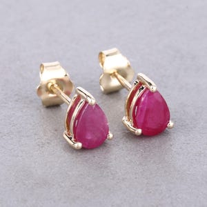 May include: A pair of gold stud earrings featuring pear-shaped, deep red gemstones. The gemstones are set in a gold frame with a post and backing for secure wear. The earrings are displayed on a neutral gray background.