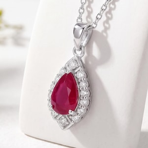 May include: A silver-toned necklace featuring a pear-shaped, deep red ruby pendant surrounded by a halo of small, clear, round-cut stones. The pendant is suspended from a delicate silver-toned chain, showcasing a polished metal setting.