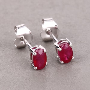 May include: A pair of silver stud earrings with oval-shaped, deep red gemstones. The gemstones are set in a four-prong setting, and the earrings have a post and butterfly backing. The earrings are displayed on a gray surface.