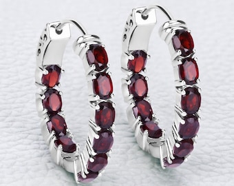 May include: A pair of silver hoop earrings, each adorned with a row of oval-cut, deep red gemstones. The earrings have a polished, reflective finish, and the gemstones are evenly spaced along the hoop's curve.