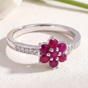 May include: A silver-toned ring featuring a flower-shaped cluster of seven round, deep pink rubies at the center. The band is accented with small, clear, round-cut stones embedded along both sides of the shank, creating a sparkling finish.