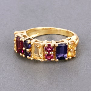 May include: Gold ring with a row of colorful gemstones. The ring features a rectangular cut amethyst, a square cut ruby, a rectangular cut citrine, a square cut ruby, and a rectangular cut amethyst.