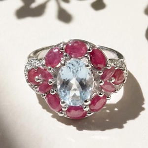 Pode incluir: Um anel de prata com uma pedra preciosa oval azul clara no centro, rodeada por pedras preciosas vermelhas ovais menores. O anel tem um design floral com pequenas pedras transparentes. O anel está sobre um fundo branco.