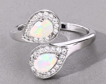 Pode incluir: Um anel de prata com duas pedras de opala em forma de pêra, cada uma rodeada por um halo de pequenas pedras preciosas transparentes. As opalas exibem um jogo de cores, com toques de verde e rosa. A banda do anel tem um acabamento liso e polido.