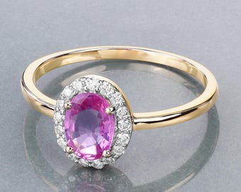 May include: A gold ring featuring an oval, pink gemstone surrounded by a halo of small, clear gemstones. The ring's band is a simple, polished gold. The ring is set against a reflective, gray surface.