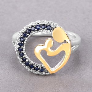 May include: A silver ring with a gold-colored mother and child design. The ring features a circle of dark blue gemstones around the mother and child. The ring is set against a gray background.